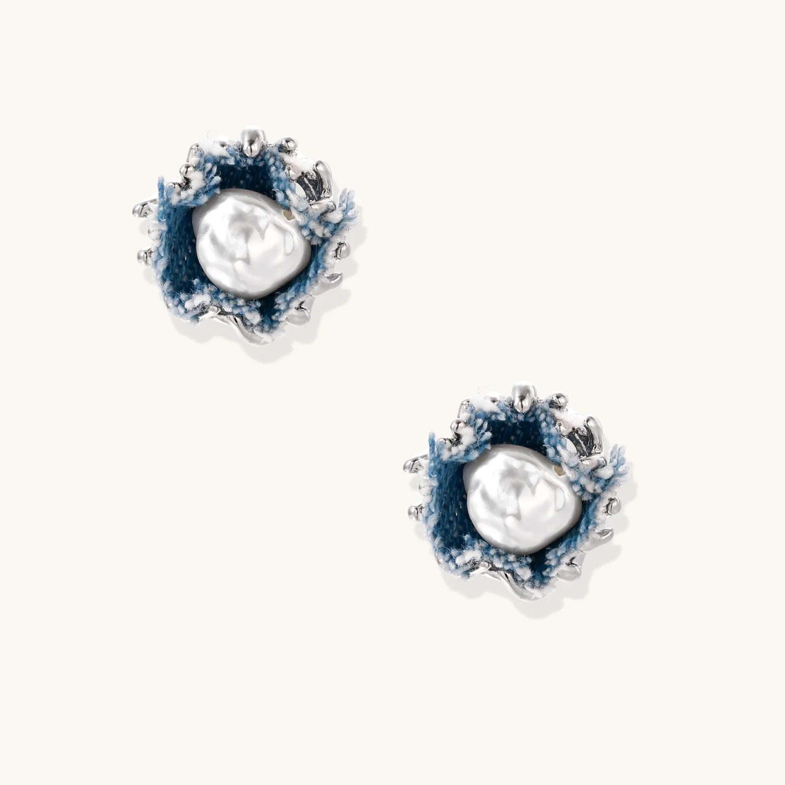 Pair of stud earrings with frayed blue denim petals around white pearl centers and silver prongs on cream background