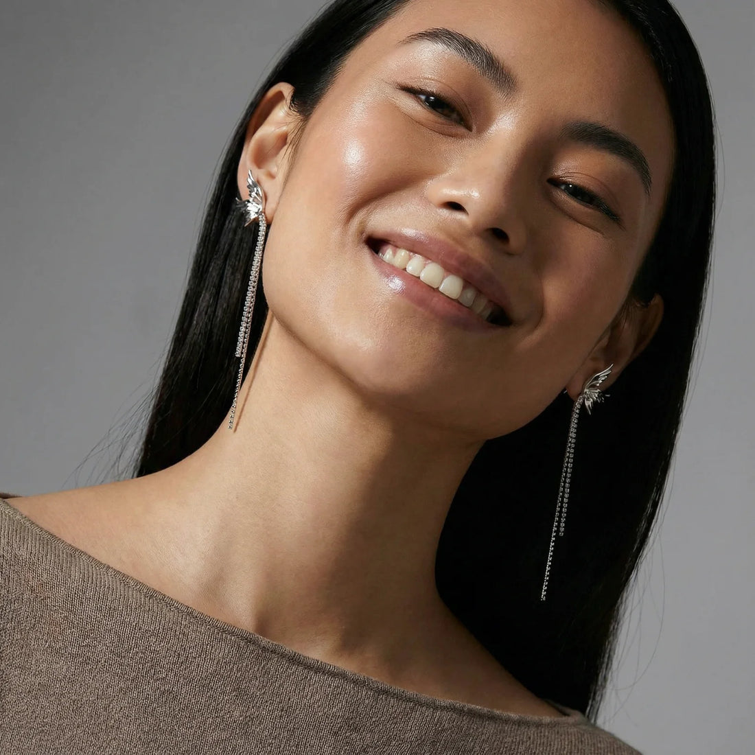 Pair of silver wing-shaped earrings with feather details and long sparkling crystal chain drops on cream background