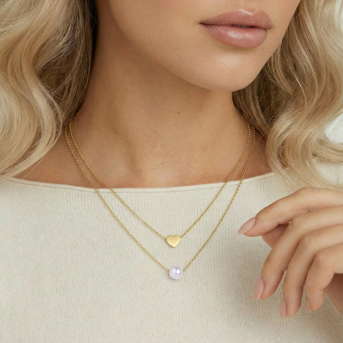 Two delicate gold-plated chains with heart charm and white pearl bead on cream background, minimal layered design.