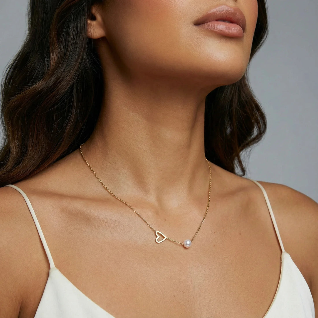 Delicate gold necklace with open heart charm and white pearl bead on fine chain against cream background
